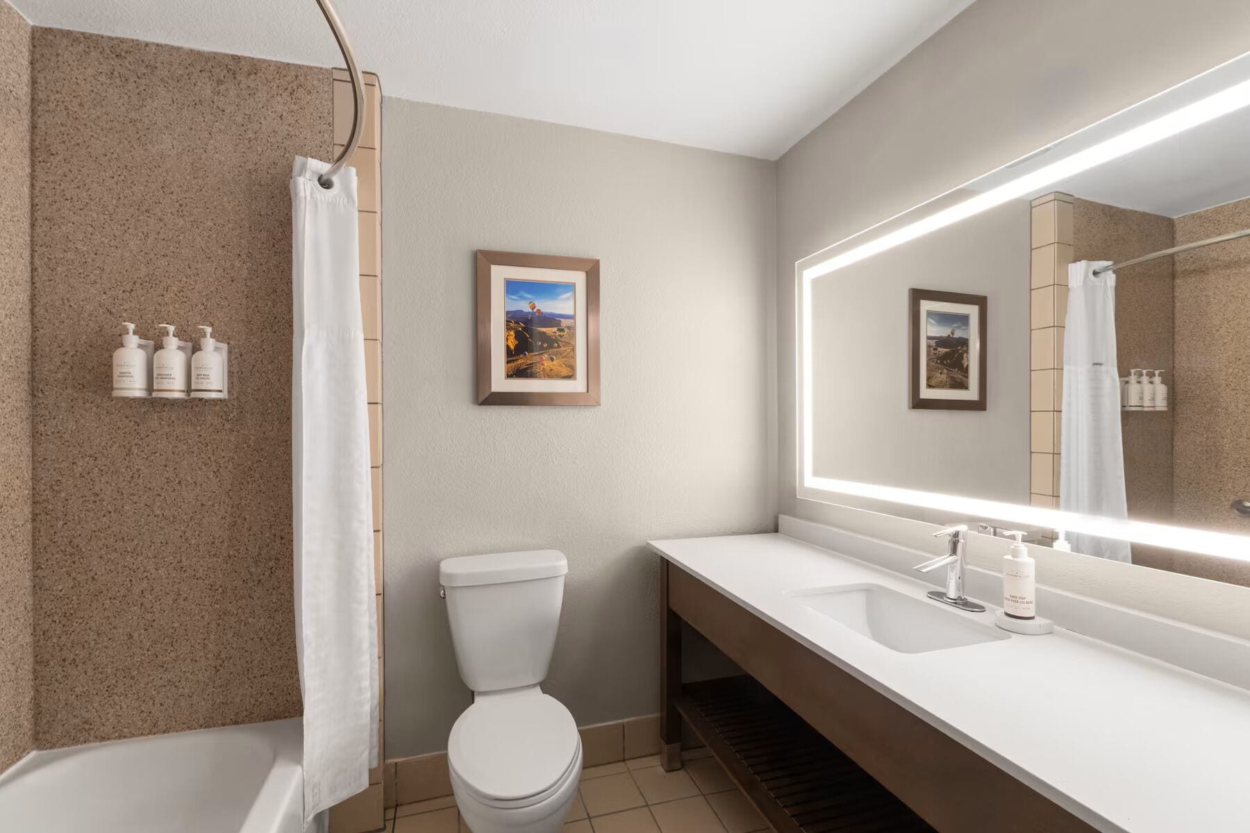 Un baño con ducha, inodoro y lavamanos, con un espejo grande y cuadros enmarcados en la pared.