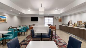 Une salle à manger d’hôtel avec plusieurs tables et chaises, une grande fenêtre avec des rideaux, une horloge au mur et un téléviseur fixé au mur.