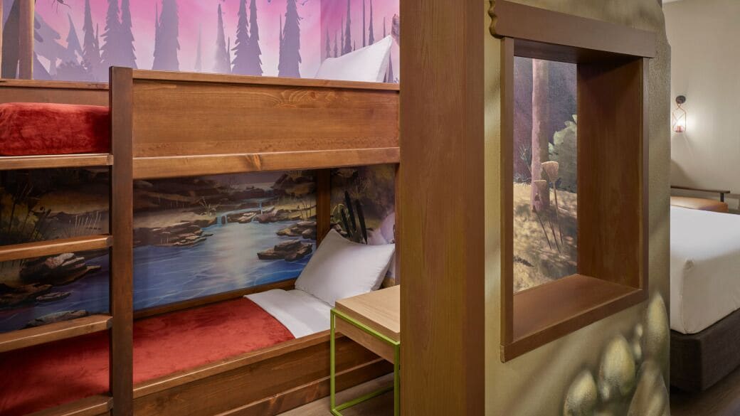Una litera con un mural de un bosque, una mesita pequeña y una ventana con vista a árboles.