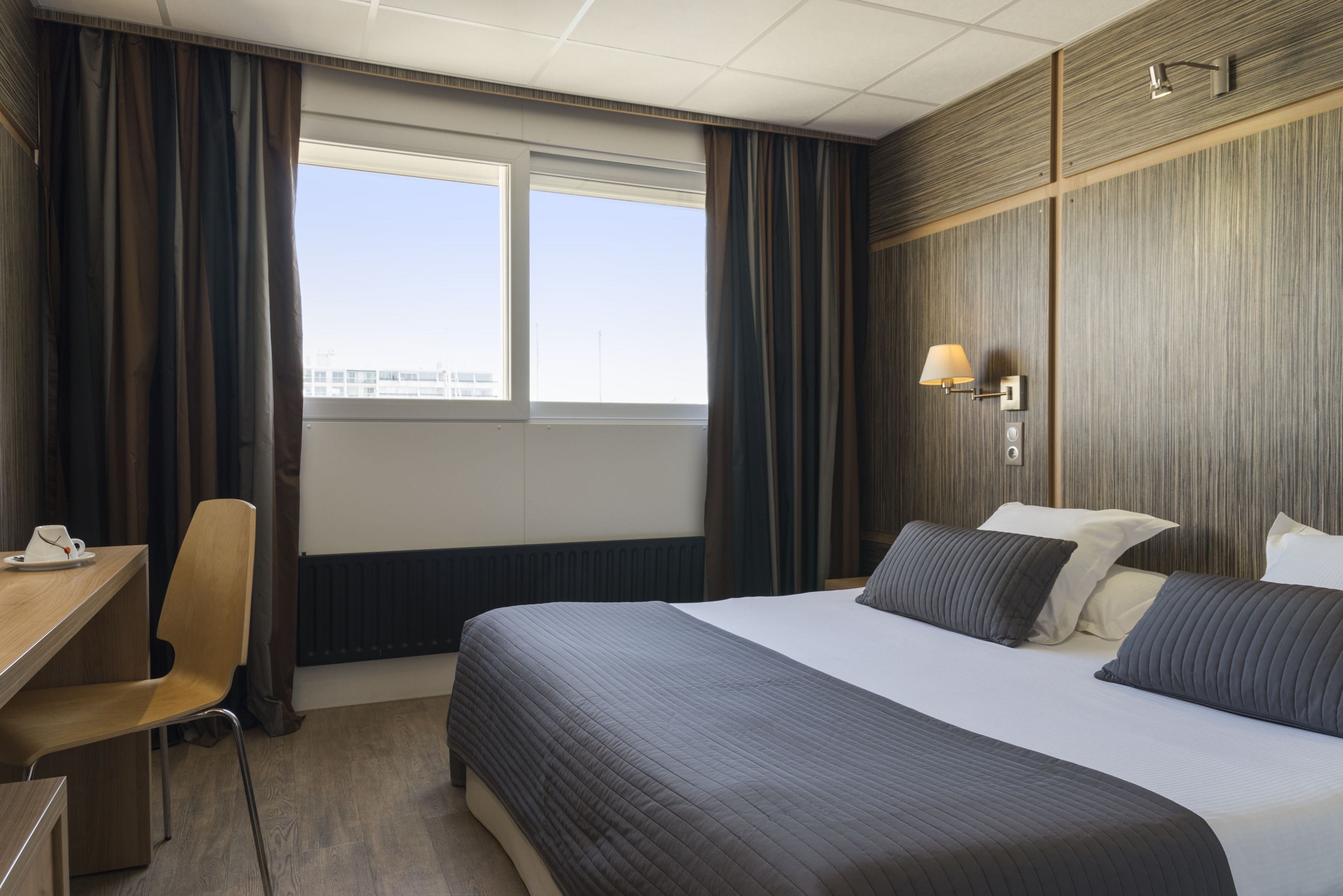 A hotel room with a large bed, a desk, a chair, and a window with curtains.