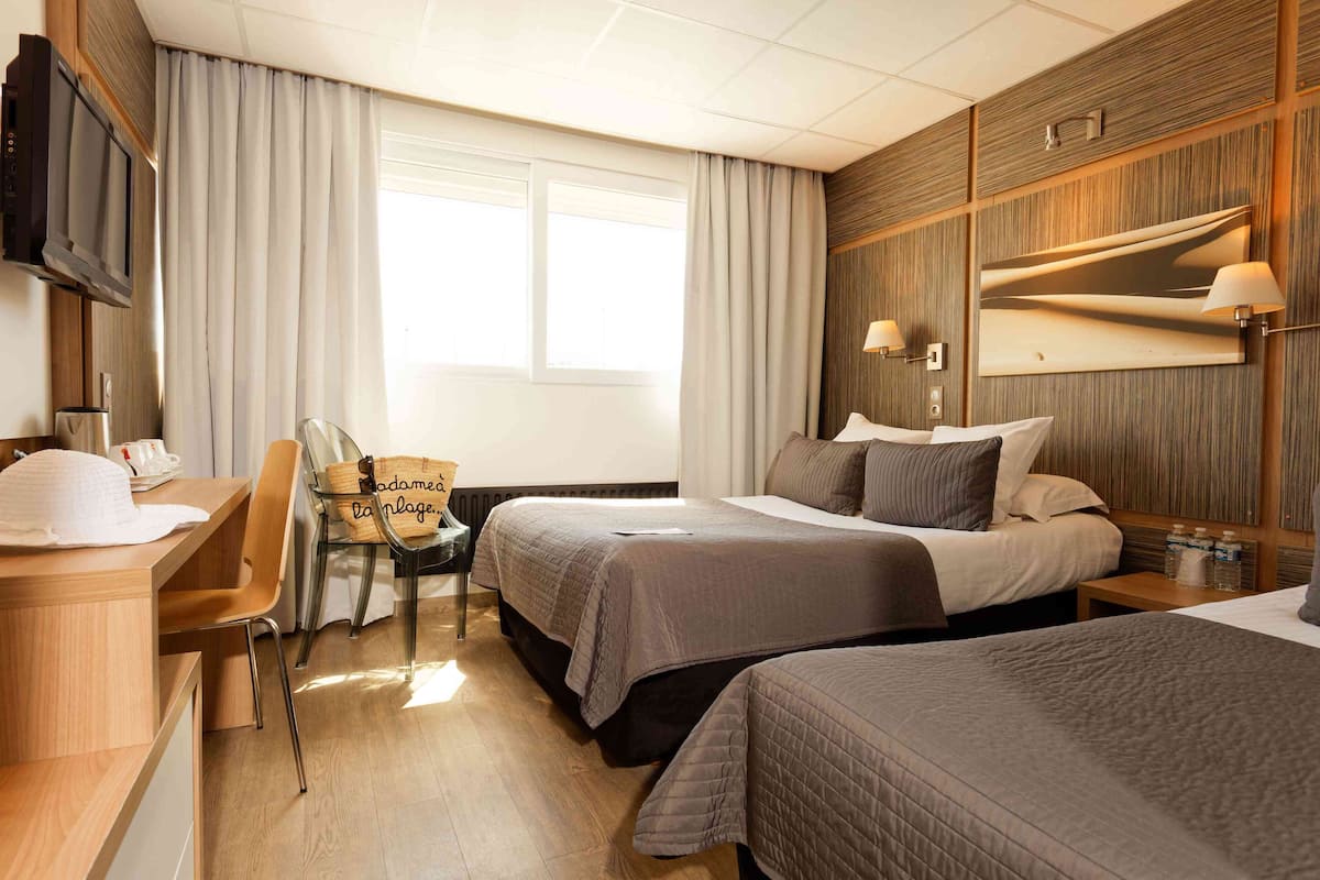Una habitación de hotel con dos camas, un escritorio con silla, televisión y un ventanal con cortinas.
