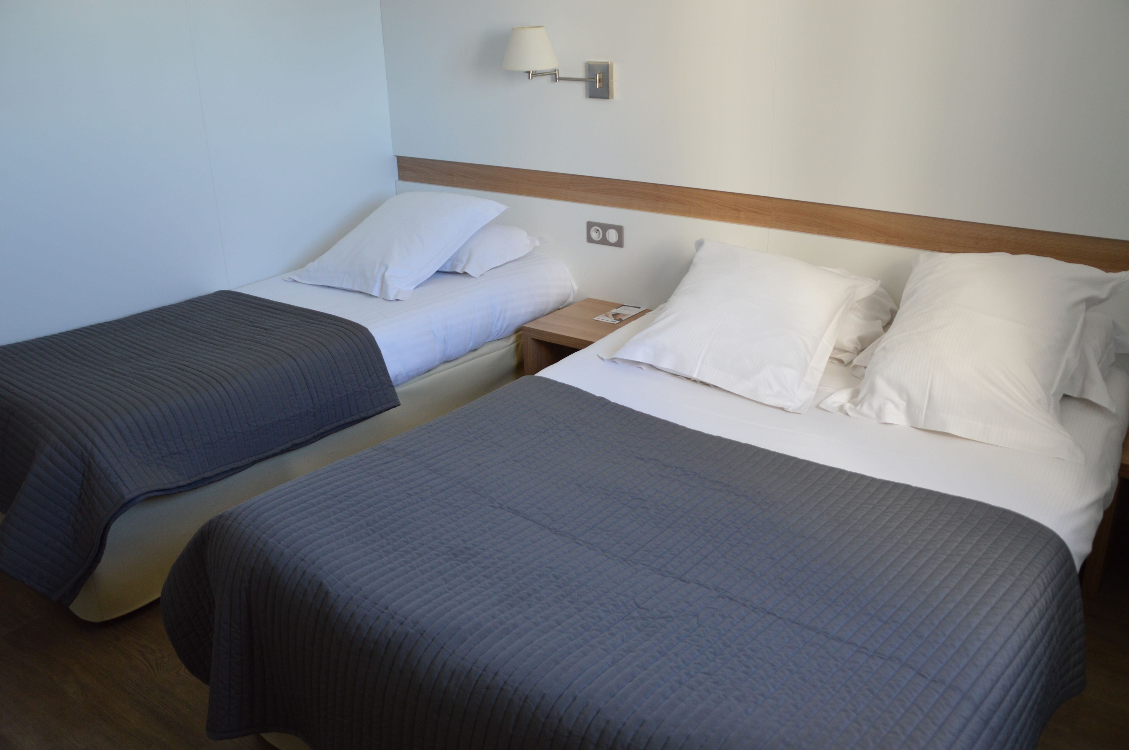 A hotel room with two single beds, each with a pillow and a blanket. There is a nightstand with a lamp and a phone.