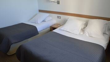 Habitación de hotel con dos camas individuales, cada una con una almohada y una manta. Hay una mesita de noche con una lámpara y un teléfono.