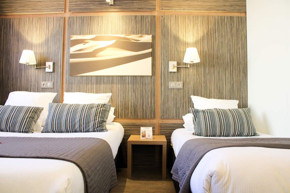 Habitación de hotel con dos camas, cada una con almohada y lámpara de noche. Hay una mesita entre las camas con un cartel encima.