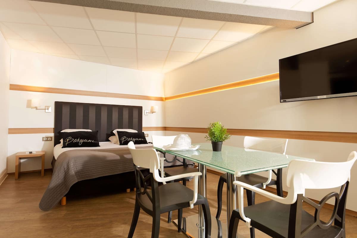 Una habitación de hotel con cama, mesitas de noche, televisor, una mesa de vidrio para comer y sillas.