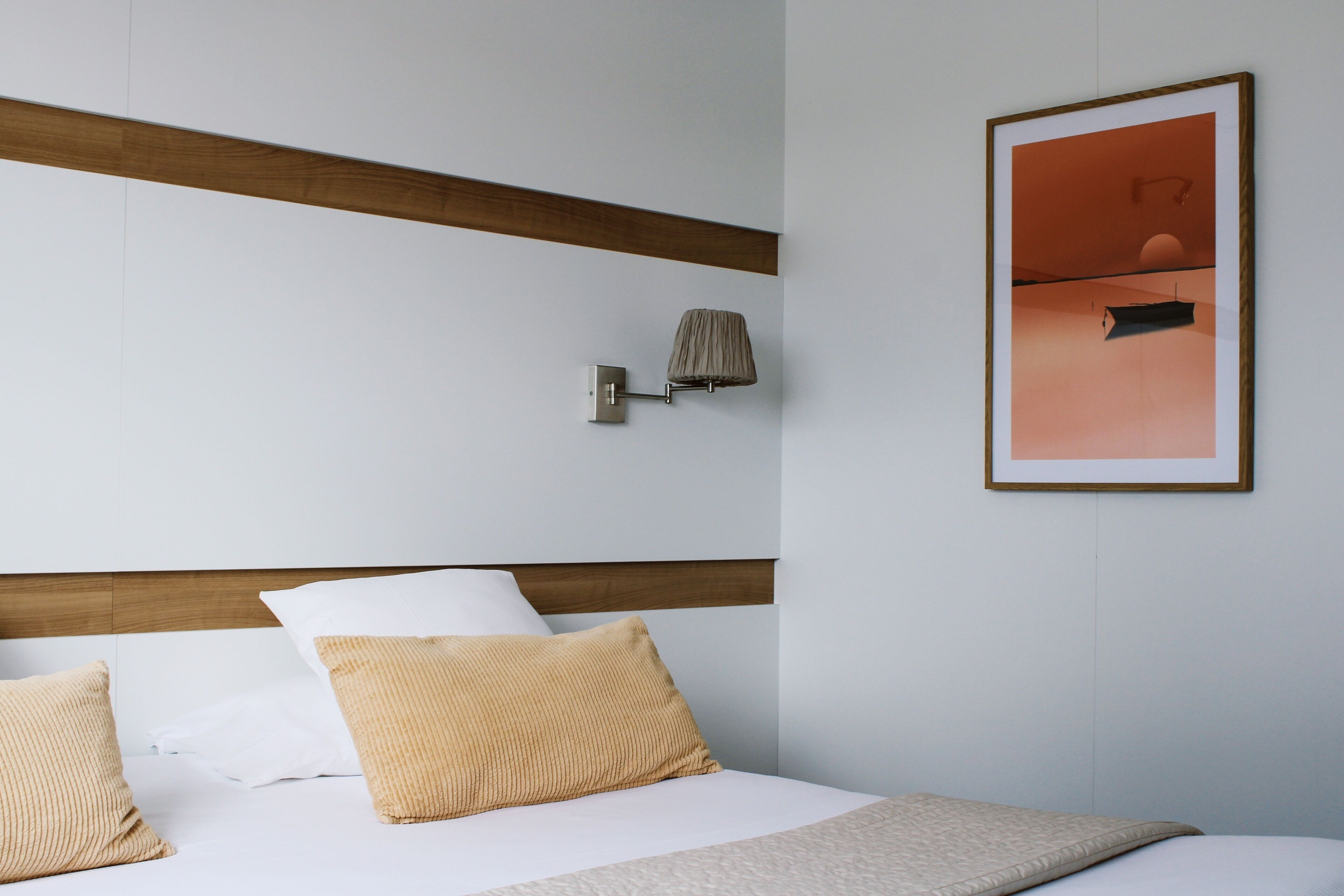 Een hotelkamer met een houten hoofdbord, een bed met wit en beige beddengoed, een wandlamp en een ingelijst beeld aan de muur.
