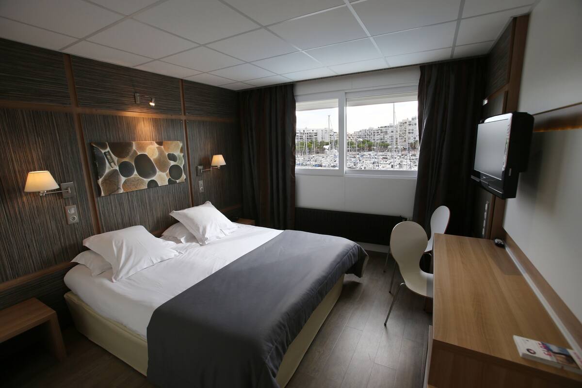 Habitación de hotel con cama, mesitas de noche, escritorio, silla, televisor y ventana con vista a barcos y edificios.
