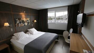 A hotel room with a bed, bedside tables, a desk, a chair, a TV, and a window with a view of boats and buildings.