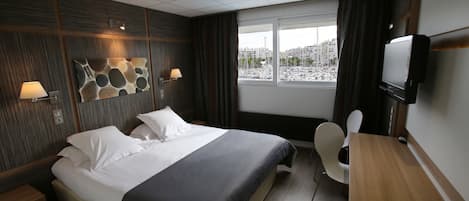 Habitación de hotel con cama, mesitas de noche, escritorio, silla, televisor y ventana con vista a barcos y edificios.