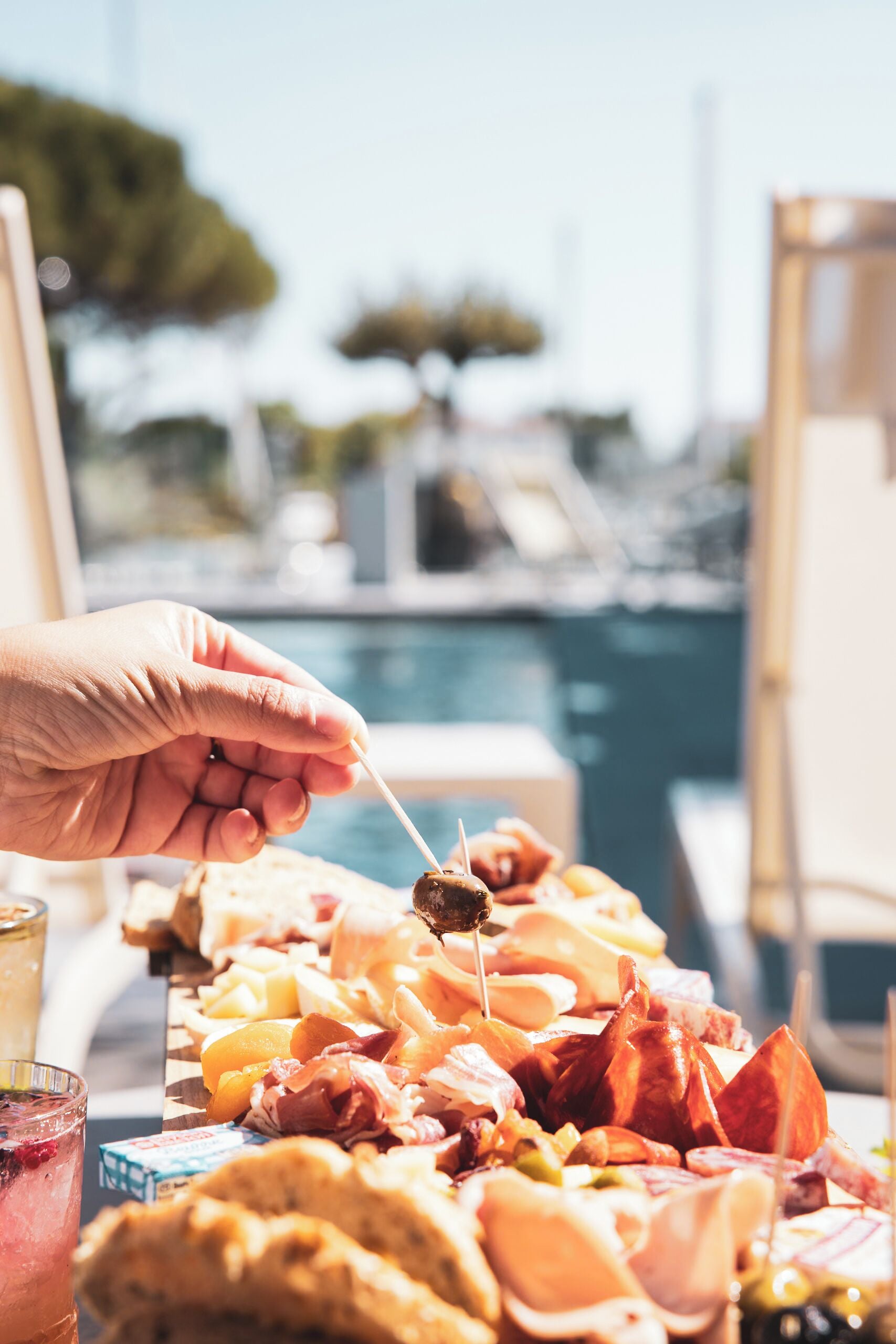 Una mano sosteniendo un pincho con comida, una mesa con diferentes platillos, y un puerto deportivo al fondo.