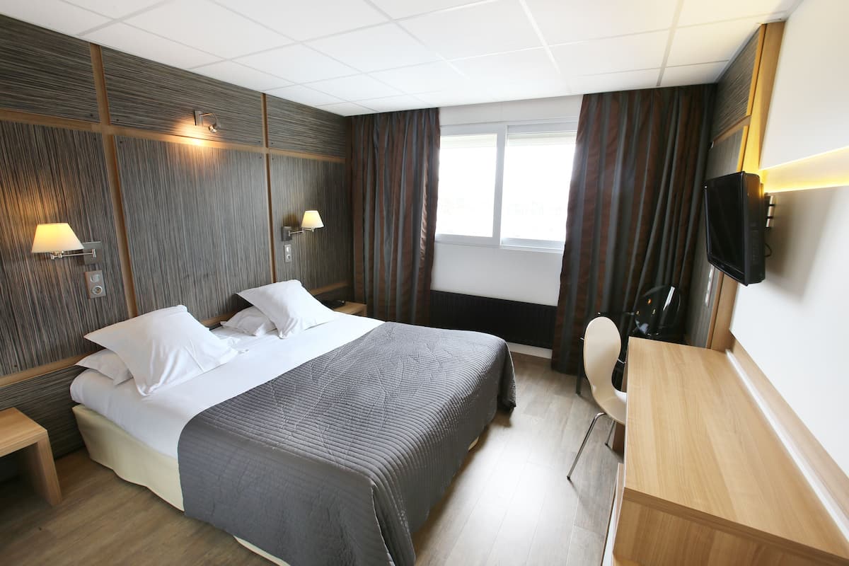 Habitación de hotel con una cama grande, mesitas de noche, un escritorio, una silla, un televisor y una ventana con cortinas.