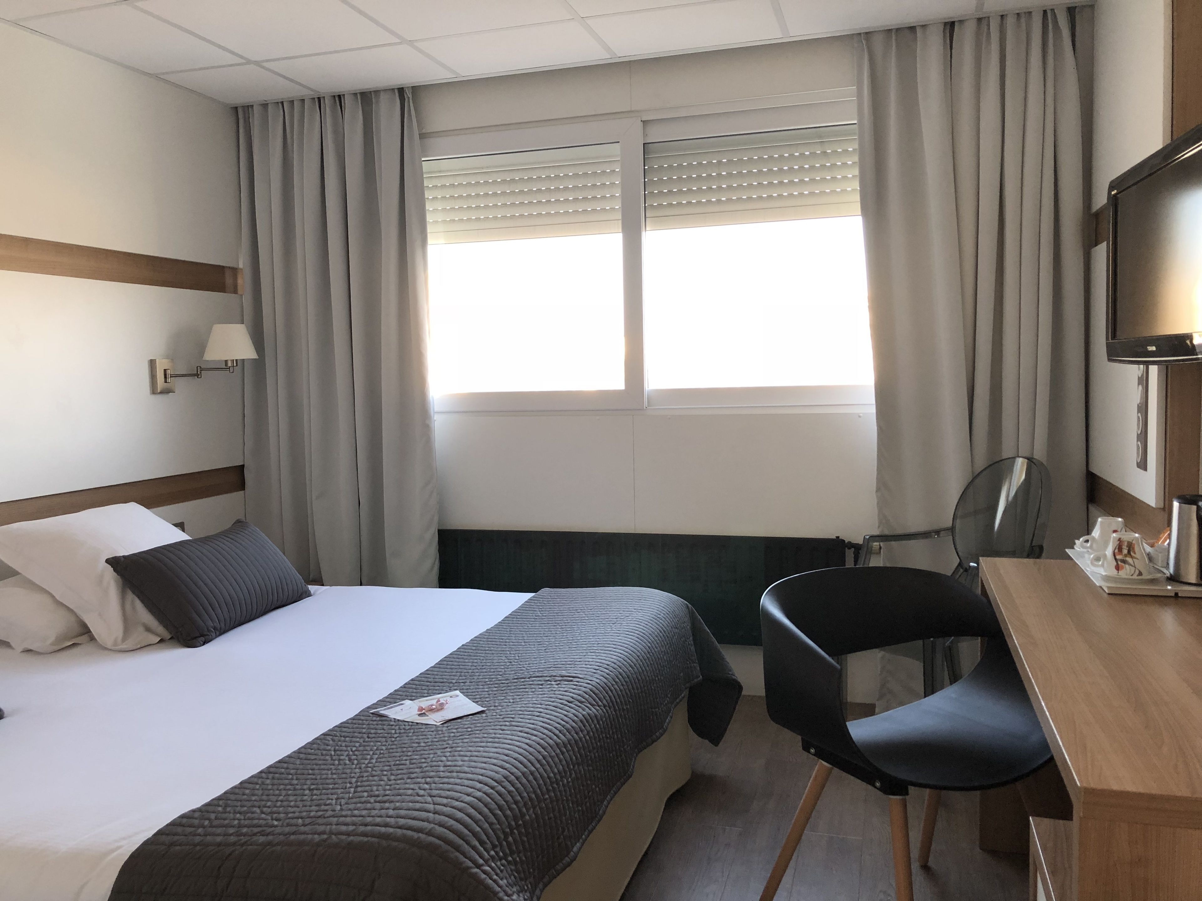 A hotel room with a bed, a desk, a chair, a TV, and a window with curtains.