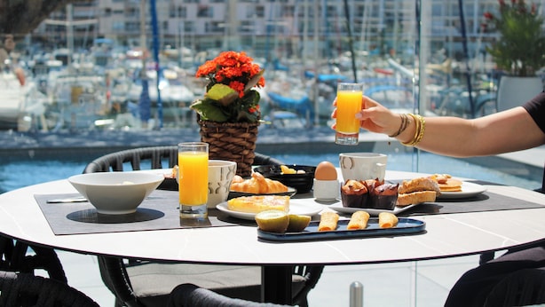 A dining table with breakfast items, a glass of orange juice, and a view of a marina with boats.