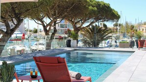 A swimming pool with lounge chairs and a table, surrounded by trees and a marina with boats.