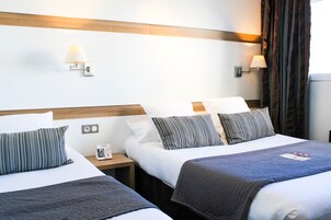 Habitación de hotel con dos camas, cada una con almohada a rayas y manta azul. Hay dos lámparas fijadas a la pared y una mesita de noche con una lámpara y un objeto pequeño encima.