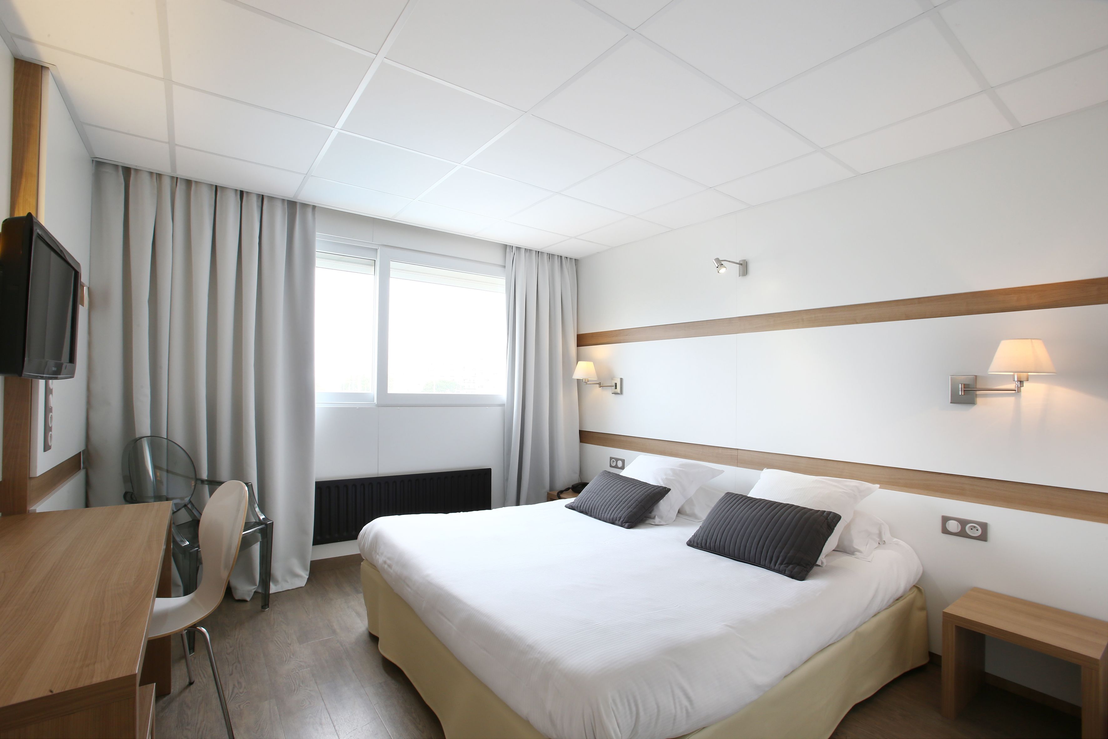 A hotel room with a bed, bedside tables, a desk, a chair, a TV, and a window with curtains.