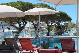 Zona de piscina con sillones de descanso y sombrillas, con vistas a un puerto deportivo con barcos.