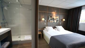Ein modernes Hotelzimmer mit einer gläsernen Dusche, einem Holz-Waschtisch, einem Bett mit dunkel gefarbener Bettdecke und einer Wandleuchte.