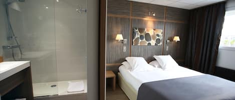 A modern hotel room with a glass-enclosed shower, a wooden vanity, a bed with a dark-colored bedspread, and a wall-mounted light fixture.