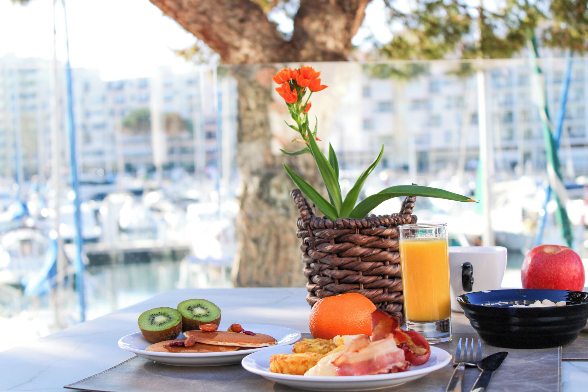 Una mesa de desayuno con platos de comida, un vaso de jugo de naranja y una cesta de mimbre con flores.