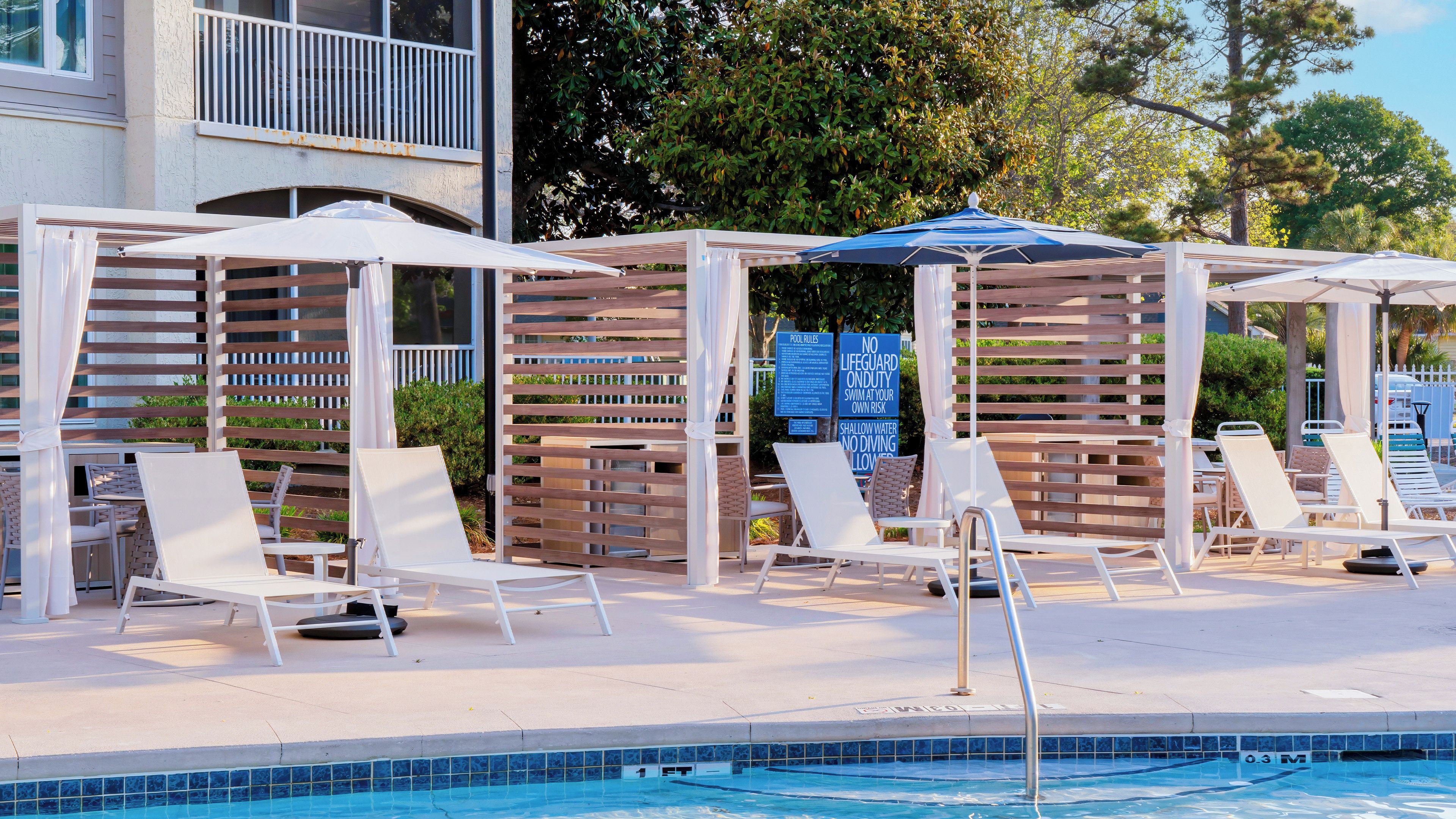 A poolside area with lounge chairs and umbrellas, a pool with a tiled edge, and a wooden pergola structure.