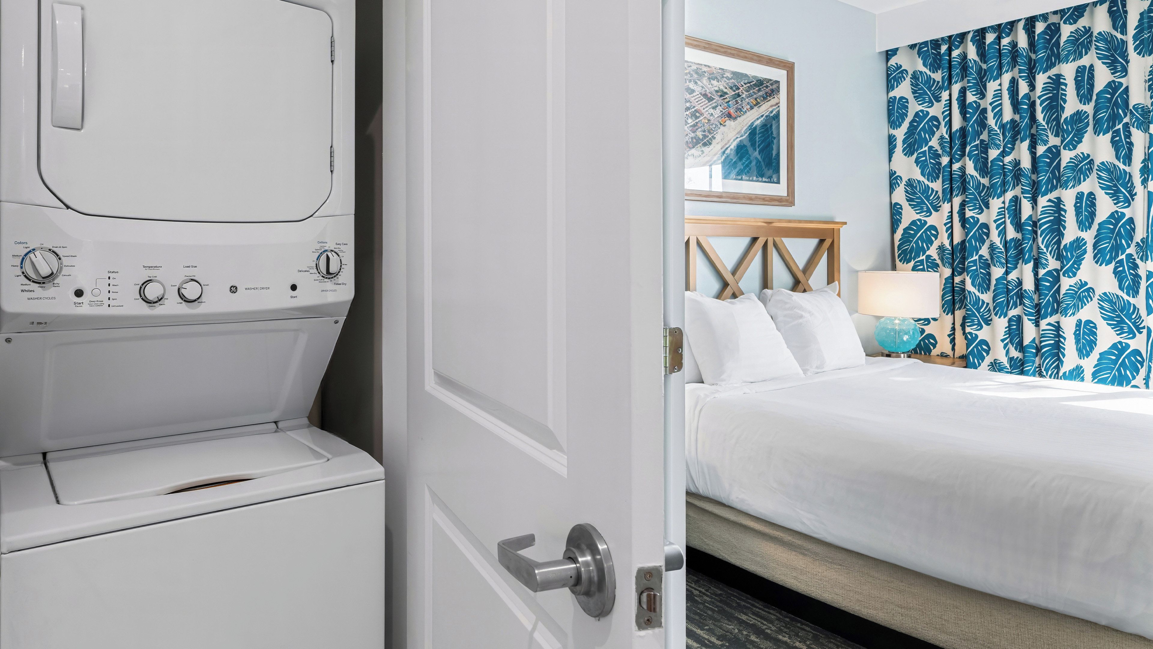 Habitación de hotel con cama, mesita de noche, lámpara, un cuadro en la pared y una lavadora.