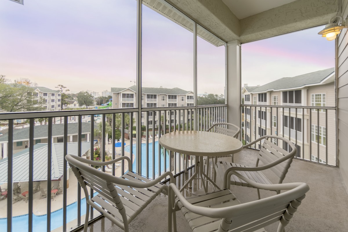 A balcony with a table and chairs, overlooking a swimming pool and residential buildings.