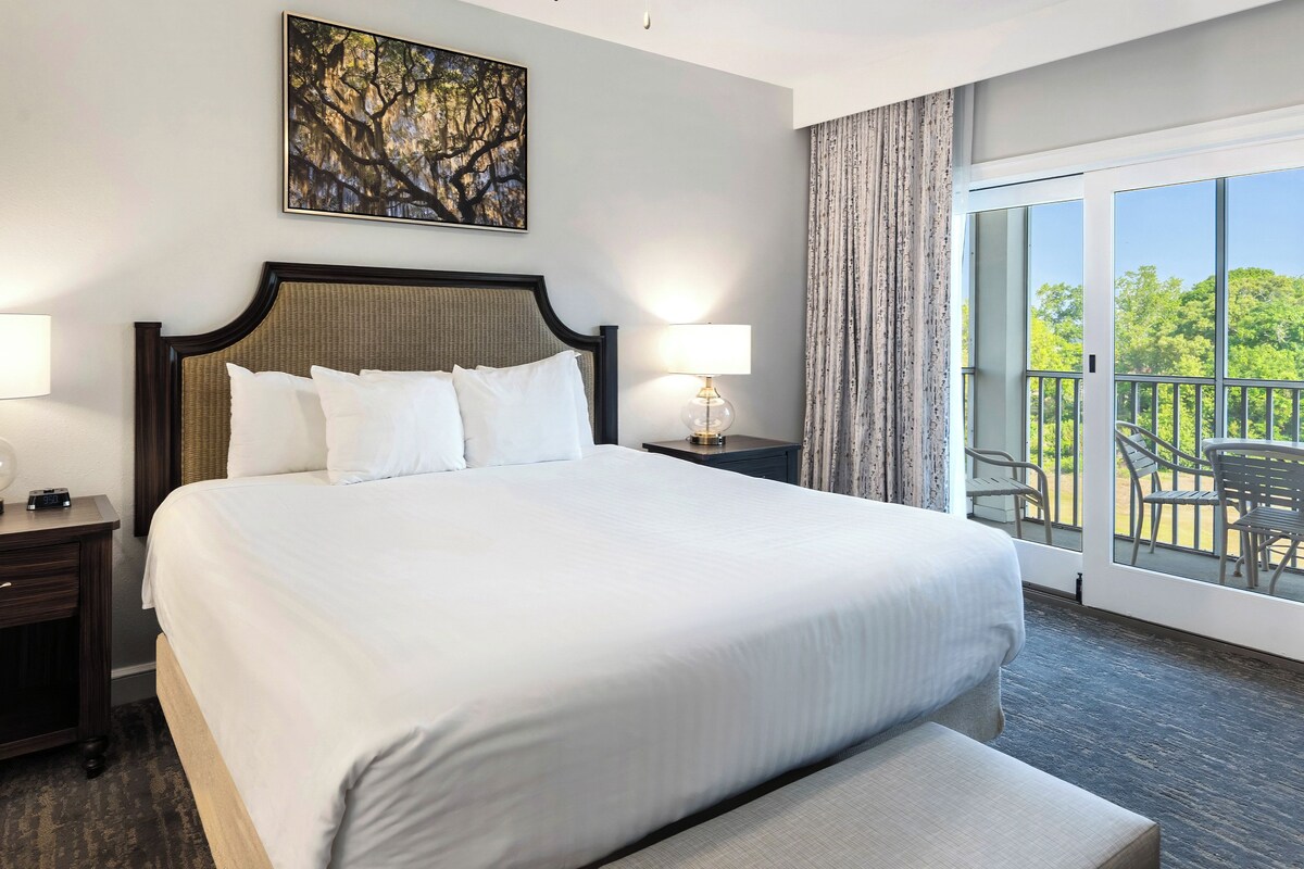 A hotel room with a large bed, two bedside tables with lamps, a painting on the wall, and a balcony with chairs.