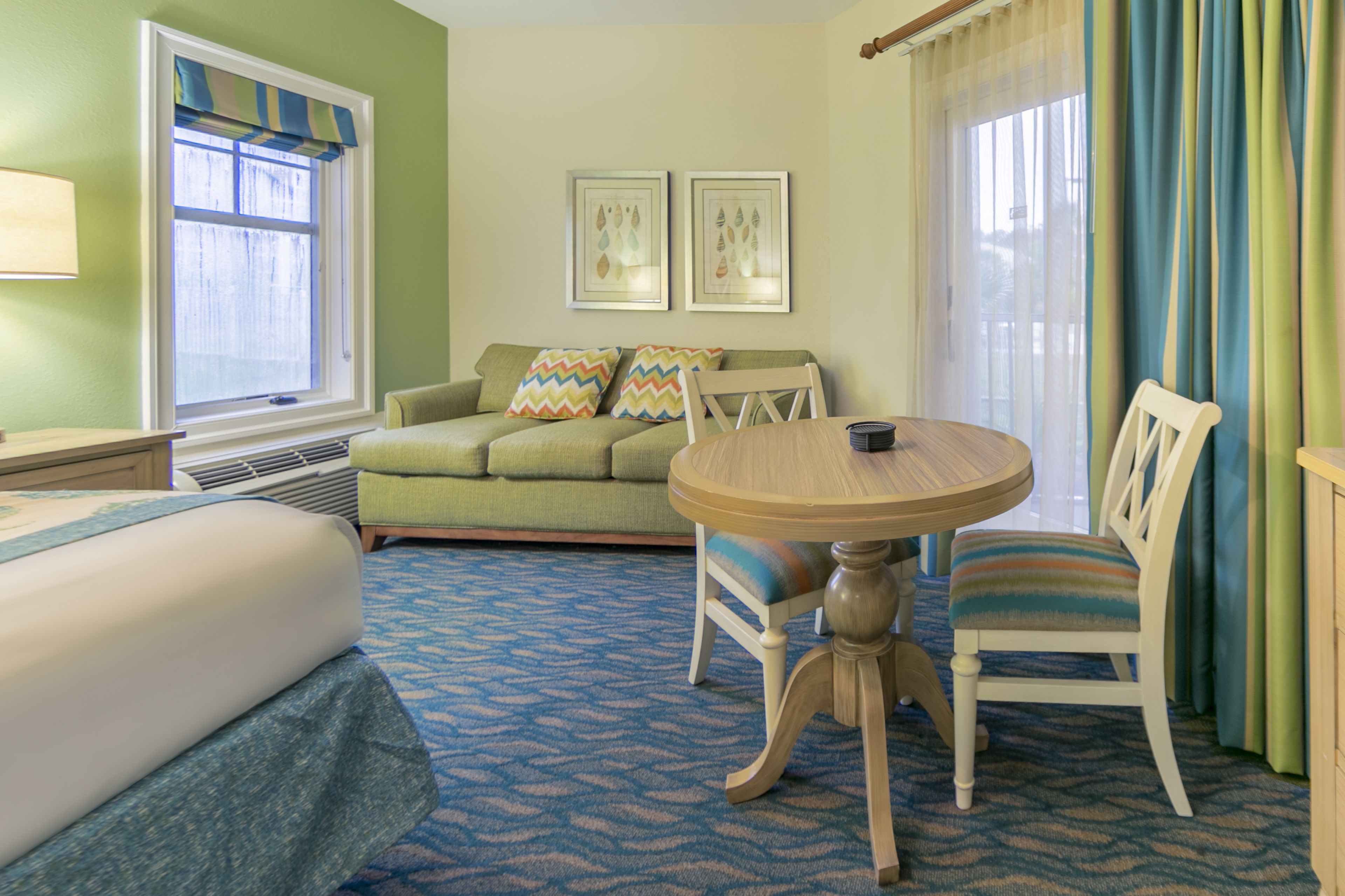 A hotel room with a bed, a green sofa, a dining table with chairs, and two framed pictures on the wall.