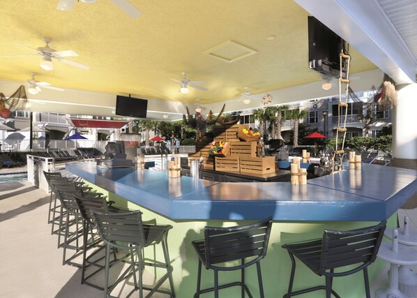 A poolside bar area with a blue counter, black bar stools, ceiling fans, and a view of the pool and palm trees in the background. - Holiday Inn Club Vacations South Beach Resort by IHG (Myrtle Beach)