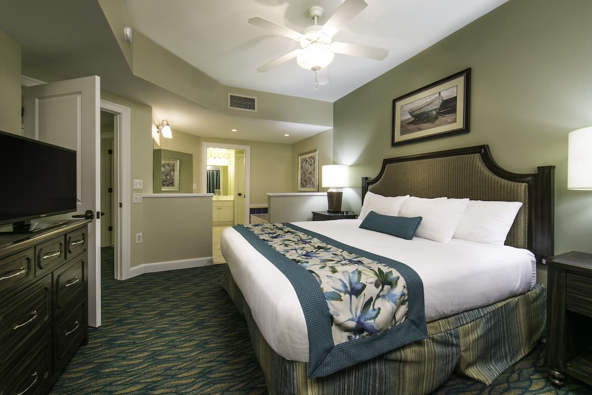 A hotel room with a bed, a television, a ceiling fan, and a painting on the wall.
