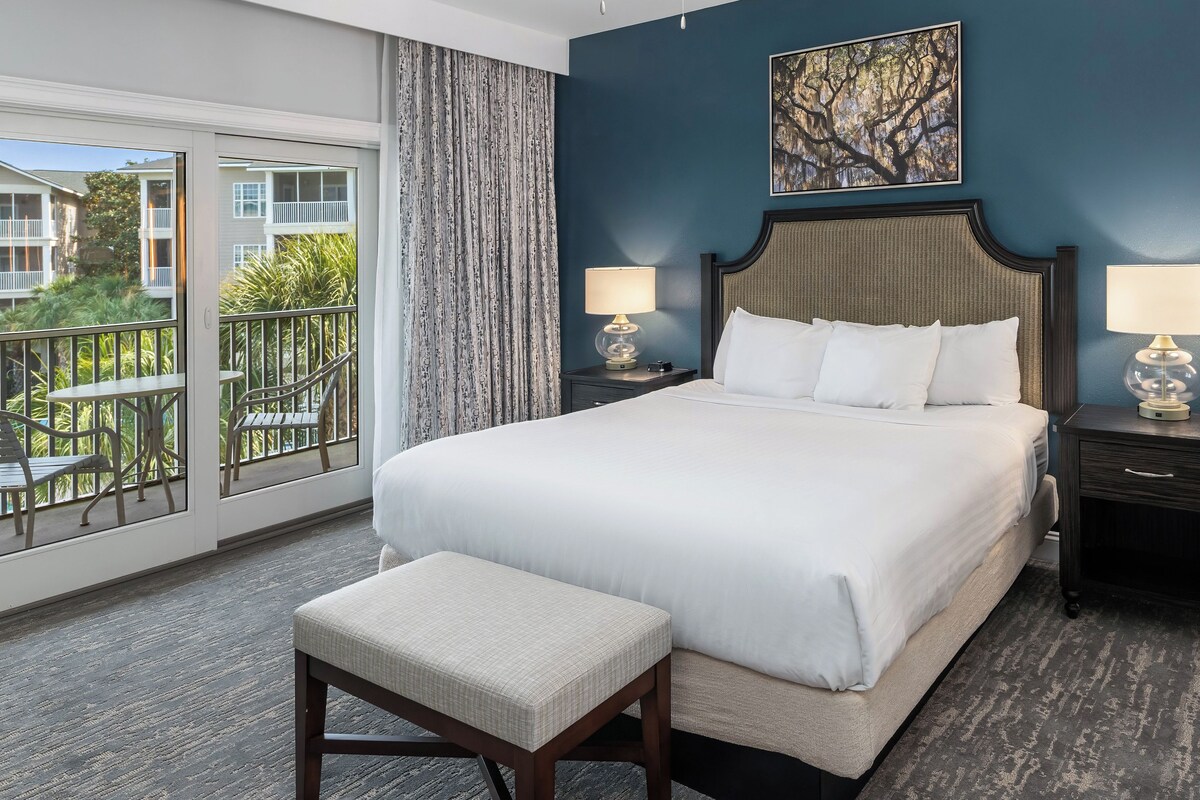A hotel room with a large bed, two bedside tables with lamps, a view of a balcony with outdoor furniture, and a painting on the wall.