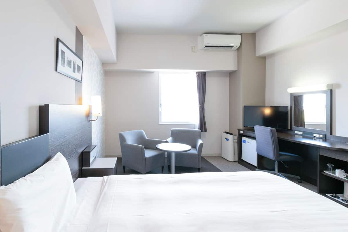 A hotel room with a bed, a desk, a chair, a TV, and an air conditioning unit.