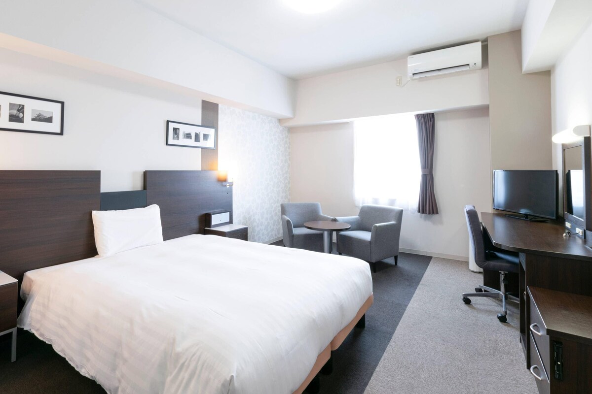 Une chambre d’hôtel avec un grand lit, un bureau avec une chaise, une petite table avec deux chaises, une télévision et un appareil de climatisation.