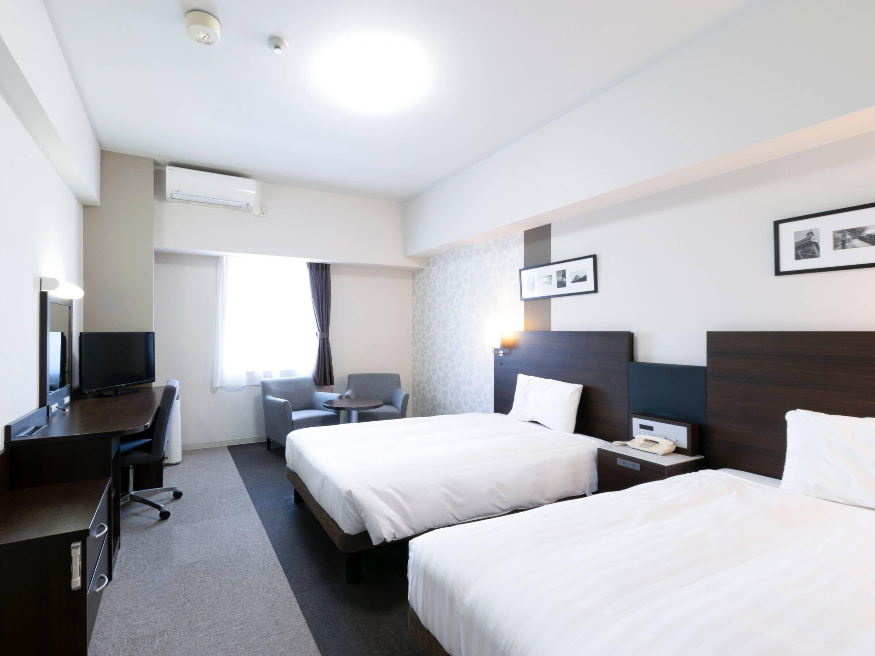 A hotel room with two beds, a desk, a chair, a TV, and a window with curtains.