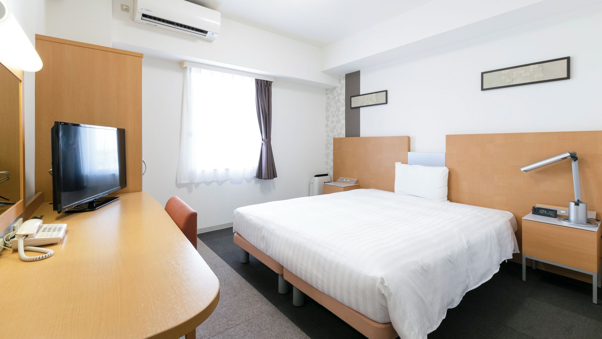 Ein Hotelzimmer mit Bett, Nachttisch, Schreibtisch mit Fernseher und Klimaanlage.