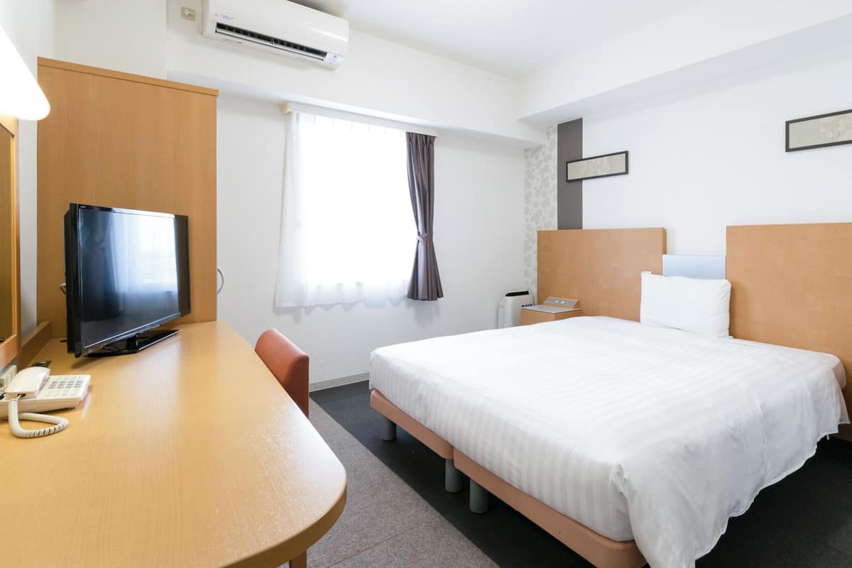 A hotel room with a bed, bedside table, desk with a TV, and an air conditioning unit.
