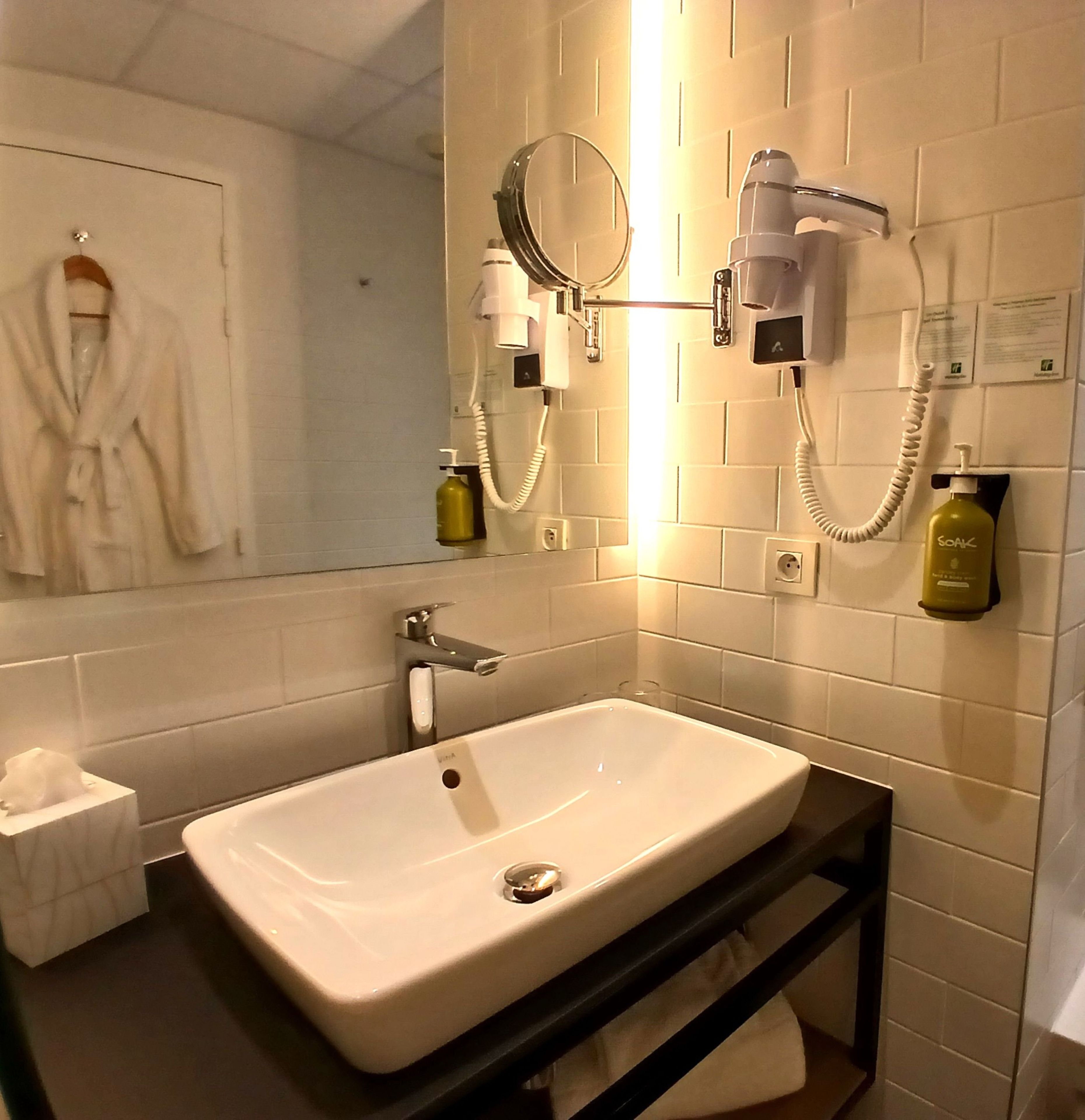 Une salle de bain avec un lavabo blanc, un miroir, un sèche-cheveux et un flacon de savon.