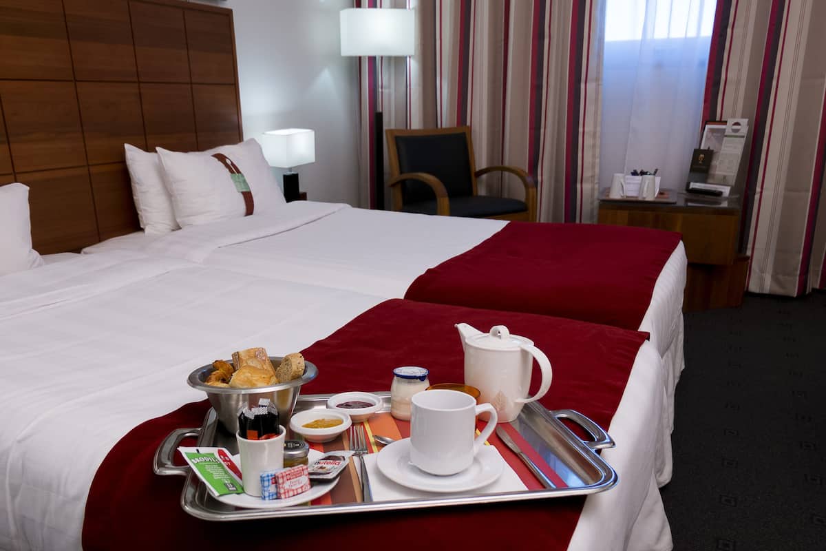 Une chambre d’hôtel avec un grand lit, un plateau contenant des articles pour le petit-déjeuner, une chaise, un bureau avec une lampe et une fenêtre avec des rideaux.