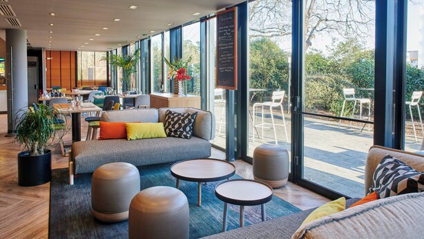 Un espace de détente intérieur moderne, doté d’un canapé, de poufs et de tables, ainsi que de grandes fenêtres offrant une vue sur l’extérieur.