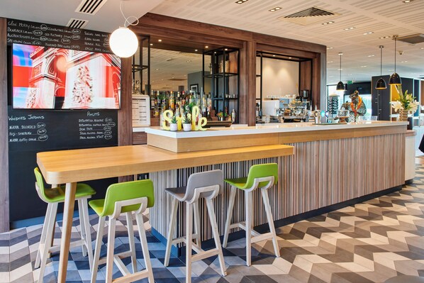 A modern bar area with a wooden counter, green and white bar stools, and a menu board displaying drink options. - Holiday Inn Bordeaux Sud - Pessac by IHG (Pessac)