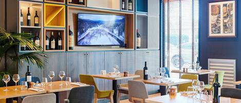 Ein Restaurant mit an der Wand montiertem Fernseher, Weinflaschen auf Holzböden und Tischen, die mit Gläsern und Besteck gedeckt sind.