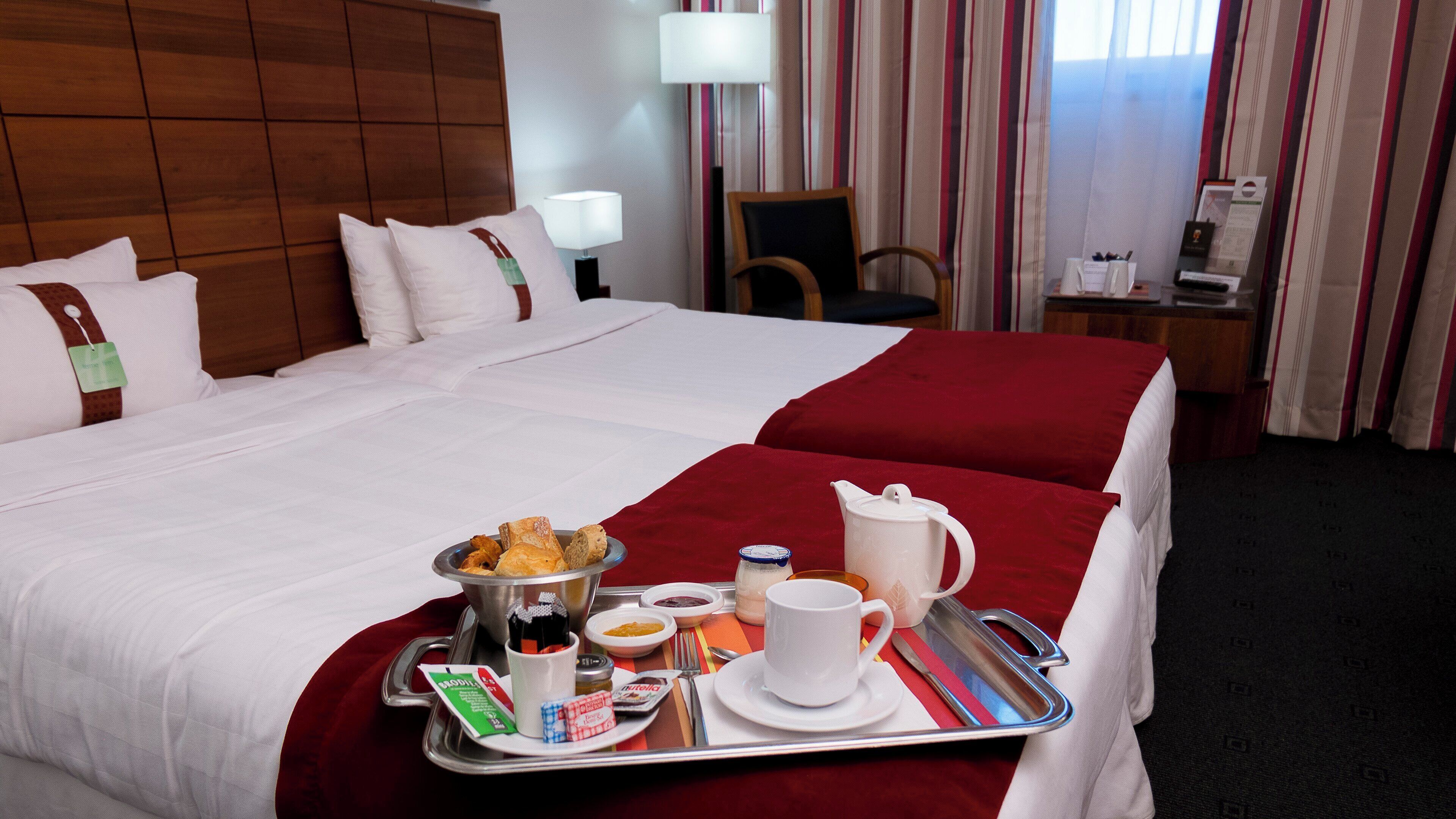 A hotel room with a large bed, a tray with breakfast items, a chair, a desk with a lamp, and a window with curtains.