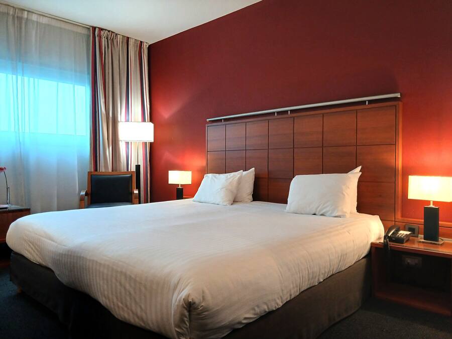 A hotel room with a large bed, two bedside tables with lamps, a wooden headboard, and a window with curtains.