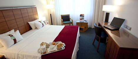Quarto de hotel com cama bem arrumada, uma escrivaninha com um laptop, uma cadeira e um tabuleiro com itens de café da manhã.