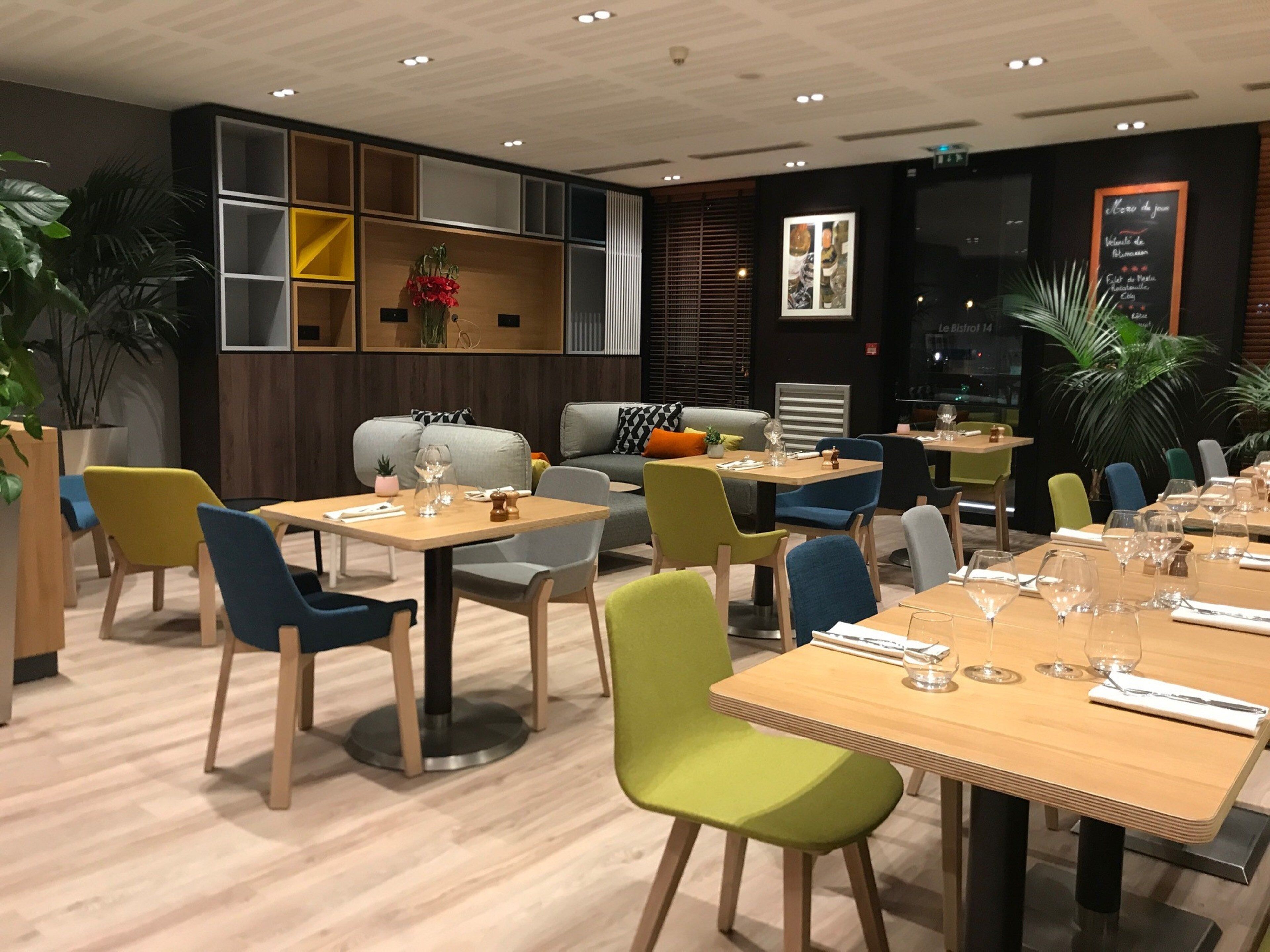 Un interior de restaurante moderno con varias mesas y sillas para comer, un sofá y plantas decorativas.