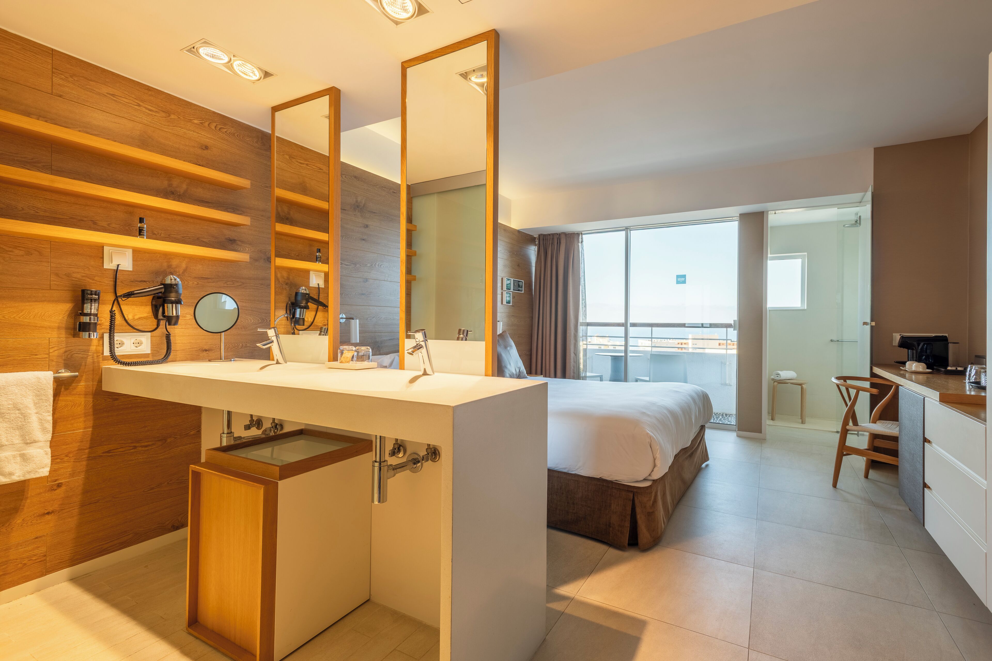 Ein modernes Hotelzimmer mit einer Wandpaneele aus Holz, einem großen Spiegel, einem weißen Waschbecken, einem Bett, einem Schreibtisch und Blick auf die Stadt.