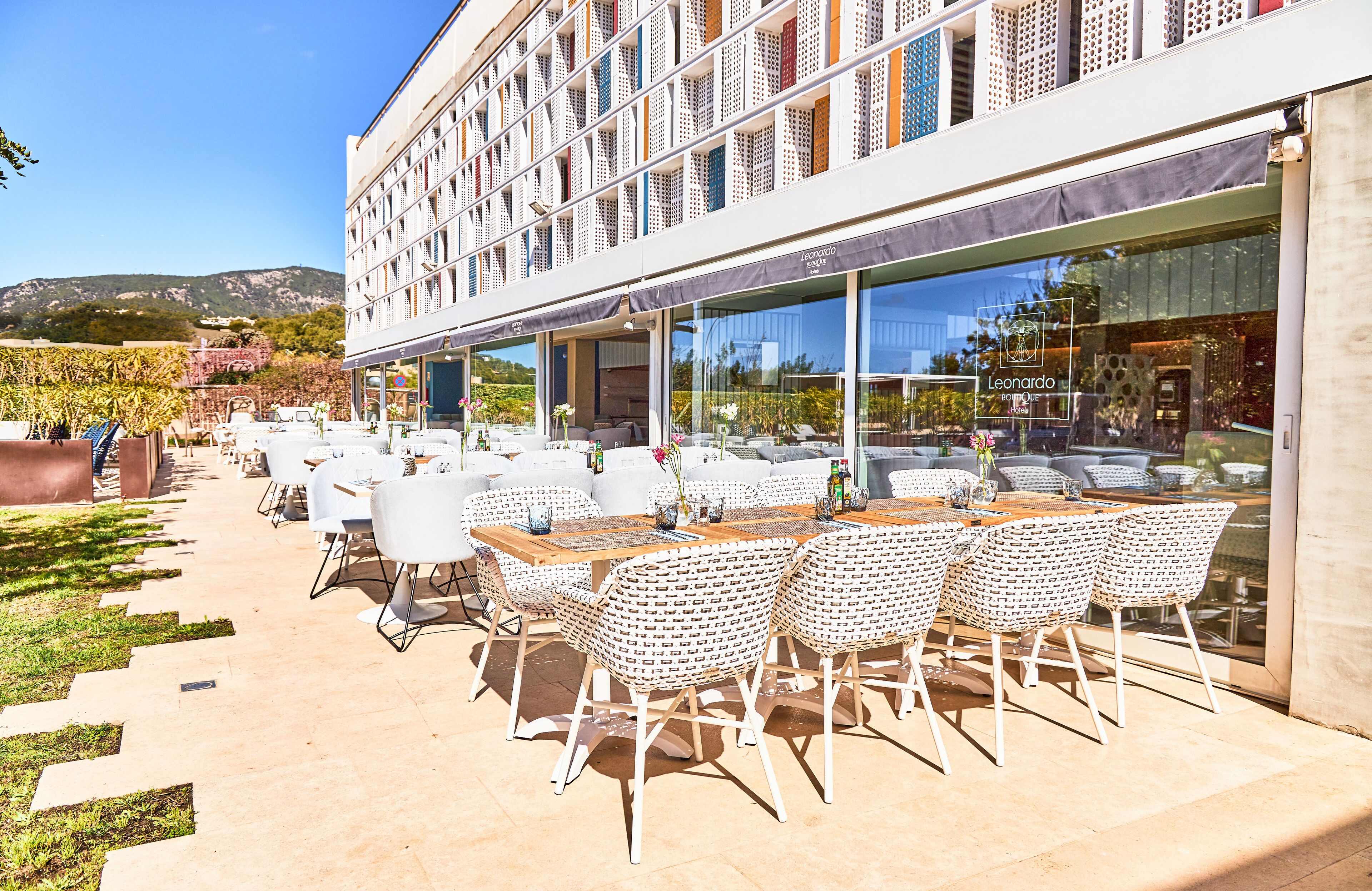 An outdoor dining area with white wicker chairs and tables, a modern building with large windows, and a view of the mountains in the background.