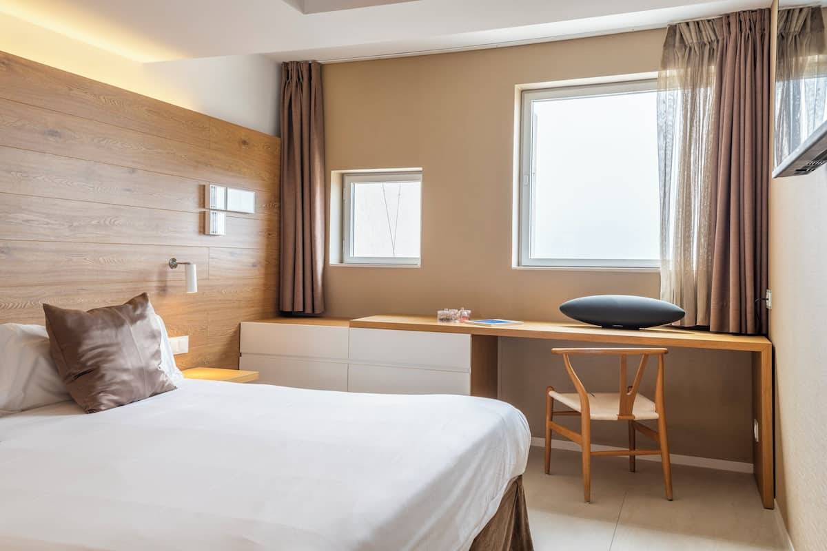 A modern hotel room with a wooden headboard, a bed with white linens, a desk with a computer, a chair, and a window with curtains.