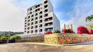 Un hotel de varios pisos con fachada de piedra y balcones, rodeado de un área ajardinada con flores y una valla.